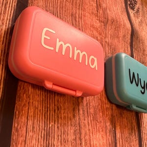 May include: Two personalized lunch boxes, one pink with the name "Emma" and one teal with the name "Wyatt".