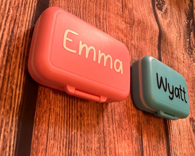 May include: Two personalized lunch boxes, one pink with the name "Emma" and one teal with the name "Wyatt".