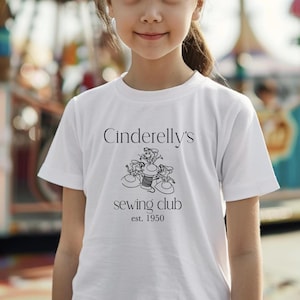 Puede incluir: Camiseta blanca con texto negro que dice "Cinderelly's sewing club est. 1950". Una ilustración en blanco y negro de ratones cosiendo está por encima del texto.