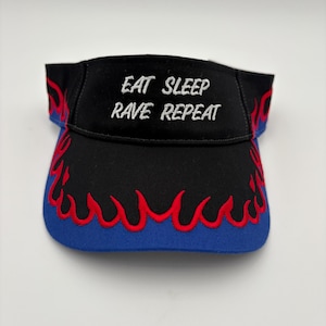 Puede incluir: Visera negra con el texto bordado "EAT SLEEP RAVE REPEAT" en blanco. La visera tiene un borde azul y detalles de llamas rojas. Un accesorio elegante para eventos al aire libre.