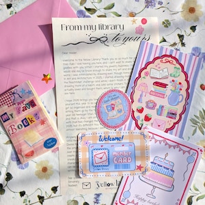 Puede incluir: Una colección de artículos de papelería, que incluye un sobre rosa, una carta, pegatinas y tarjetas de felicitación. Las tarjetas presentan ilustraciones de libros, una tarta y una tarjeta de biblioteca. Los artículos están dispuestos sobre una superficie con estampado floral.