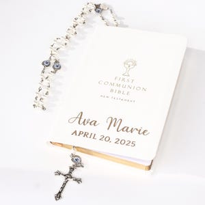 May include: A white First Communion Bible with gold lettering and the name "Ava Marie" and the date "April 20, 2025". A rosary with white beads and a silver cross are also present.