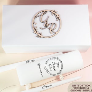 May include: White gift box with two white candles, one labeled "Bride" and the other "Groom". The candles are decorated with a floral wreath design and the text "Your Design Here". The box is decorated with a wooden circle with the initials "C & R" and a floral wreath design.