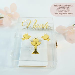 May include: A clear acrylic box with gold accents, featuring the name "Noah" in gold script. The box is decorated with gold dove and chalice designs. The text "PERSONALIZED BIBLE ACRYLIC BOX" is visible.