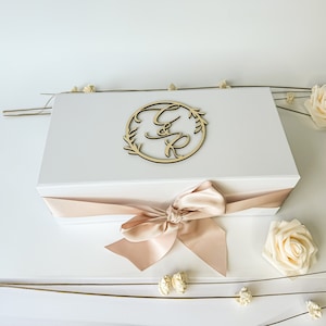 May include: A white gift box with a beige satin ribbon tied in a bow. The box has a wooden circle with the letters "C & R" in a cursive font and a leafy wreath design on top.