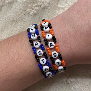 Könnte beinhalten: Zwei Perlenarmbänder mit den Buchstaben "VERPISTA" in weißen Buchstabenperlen. Ein Armband ist blau und schwarz, das andere orange und schwarz. Die Armbänder werden an einem Handgelenk getragen.