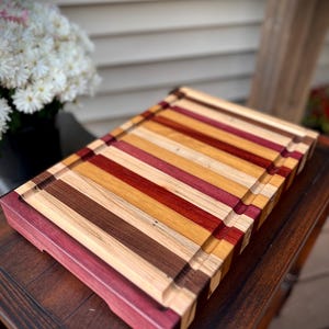 May include: A large, rectangular cutting board made of various types of wood. The board has a natural wood finish and features a variety of colors, including red, brown, and yellow. The board has a raised edge around the perimeter and a groove around the edge for catching juices.