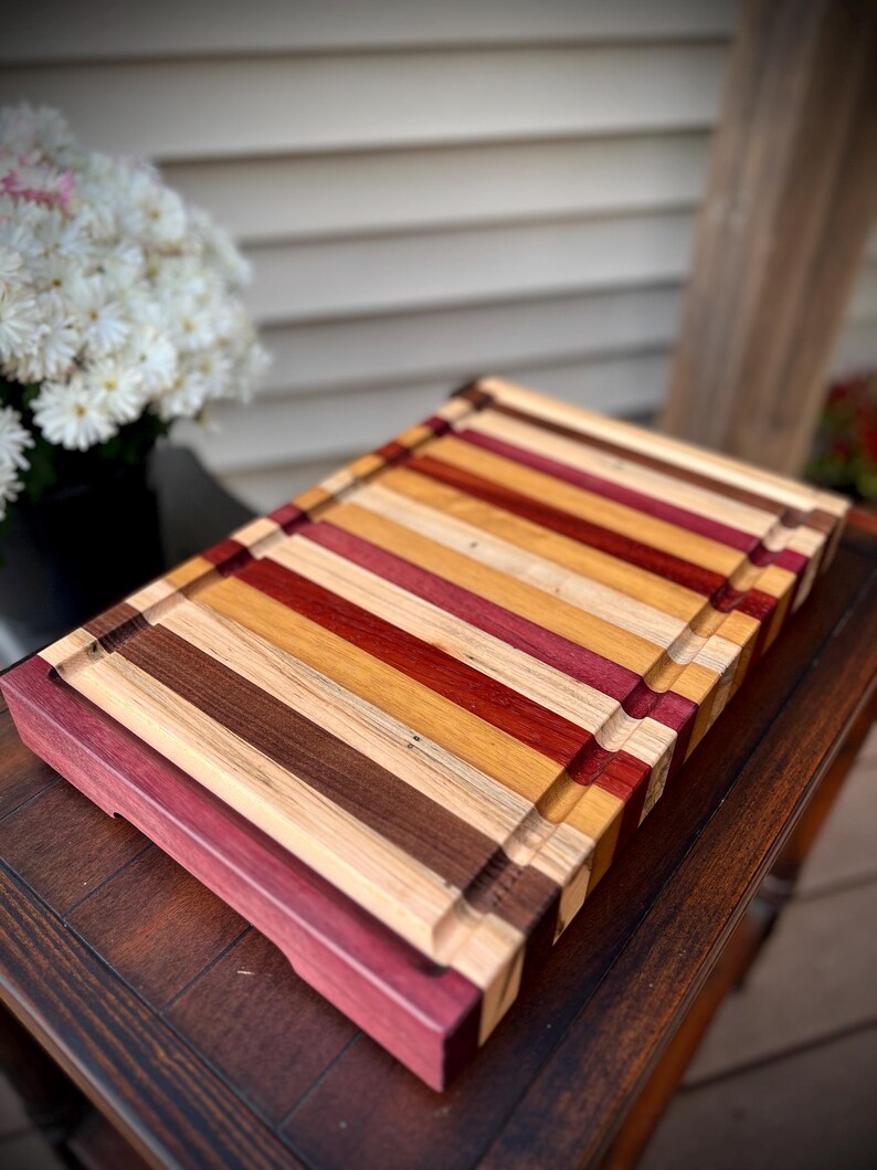May include: A large, rectangular cutting board made of various types of wood. The board has a natural wood finish and features a variety of colors, including red, brown, and yellow. The board has a raised edge around the perimeter and a groove around the edge for catching juices.