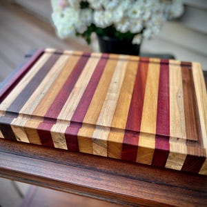 May include: A large, rectangular cutting board made of various types of wood. The board has a natural wood finish and is striped with different shades of brown, red, and purple wood.