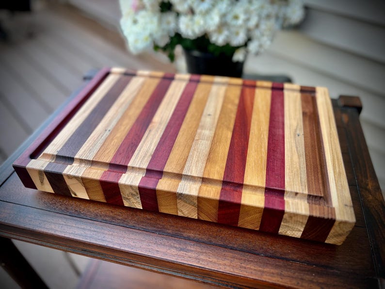 May include: A large, rectangular cutting board made of various types of wood. The board has a natural wood finish and is striped with different shades of brown, red, and purple wood.
