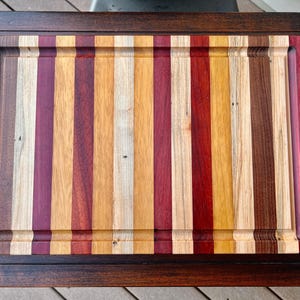 May include: A large, rectangular cutting board made of various types of wood. The board has a dark brown frame and a center section made of alternating strips of light and dark wood. The center section is surrounded by a border of darker wood.