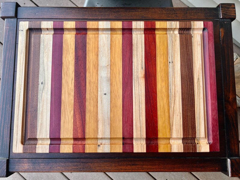 May include: A large, rectangular cutting board made of various types of wood. The board has a dark brown frame and a center section made of alternating strips of light and dark wood. The center section is surrounded by a border of darker wood.