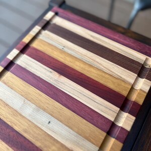 May include: A large wooden cutting board with a dark wood frame. The board is made of multiple strips of wood in various shades of brown, red, and yellow.