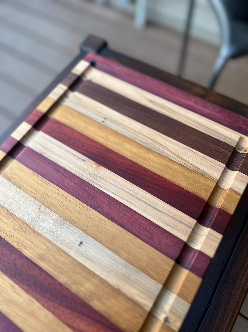 May include: A large wooden cutting board with a dark wood frame. The board is made of multiple strips of wood in various shades of brown, red, and yellow.