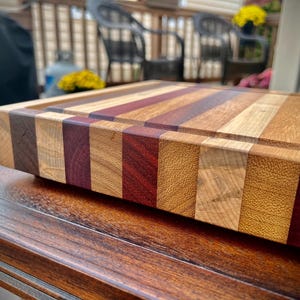 May include: A large, rectangular wooden cutting board with a multi-colored striped design. The board is made of different types of wood, including maple, walnut, and cherry. The board has a natural finish and is perfect for cutting and serving food.