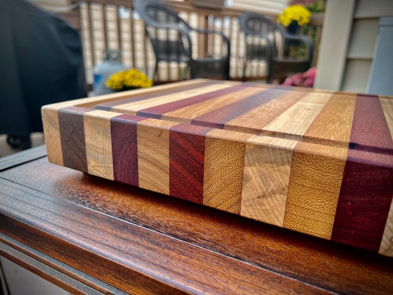 May include: A large, rectangular wooden cutting board with a multi-colored striped design. The board is made of different types of wood, including maple, walnut, and cherry. The board has a natural finish and is perfect for cutting and serving food.