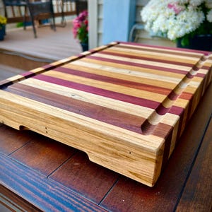 May include: A large, rectangular wooden cutting board with a natural finish. The board is made of multiple strips of wood, each with a different color and grain pattern. The board has a raised edge around the perimeter and a groove running along the length of the board.