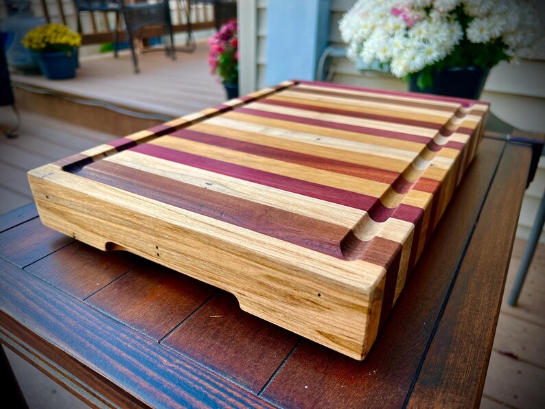 May include: A large, rectangular wooden cutting board with a natural finish. The board is made of multiple strips of wood, each with a different color and grain pattern. The board has a raised edge around the perimeter and a groove running along the length of the board.