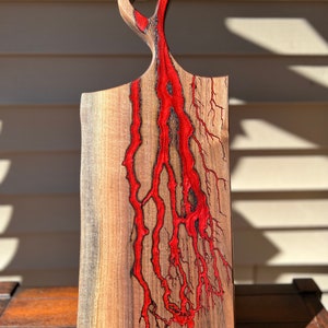 Op de afbeelding: Een houten snijplank met een uniek, abstract ontwerp. De plank is gemaakt van walnoot hout en heeft een rode epoxyhars die door het hout stroomt. De plank heeft een handvat aan de bovenkant en is perfect voor het serveren van kaas, charcuterie of andere hapjes.