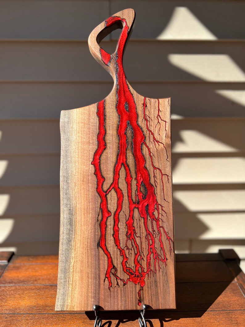 Op de afbeelding: Een houten snijplank met een uniek, abstract ontwerp. De plank is gemaakt van walnoot hout en heeft een rode epoxyhars die door het hout stroomt. De plank heeft een handvat aan de bovenkant en is perfect voor het serveren van kaas, charcuterie of andere hapjes.