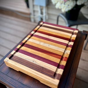 May include: A large, rectangular wooden cutting board with a natural finish. The board is made of multiple strips of wood in various shades of brown, red, and yellow, creating a striped pattern. The board has a raised edge around the perimeter.