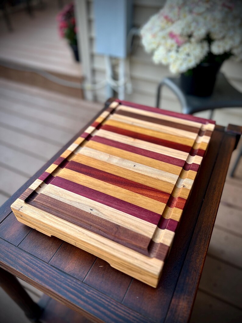 May include: A large, rectangular wooden cutting board with a natural finish. The board is made of multiple strips of wood in various shades of brown, red, and yellow, creating a striped pattern. The board has a raised edge around the perimeter.