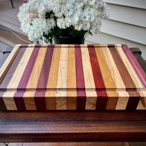May include: A large, rectangular cutting board made of various types of wood. The board has a striped pattern of light and dark wood, with a darker wood border. The board is sitting on a wooden table.