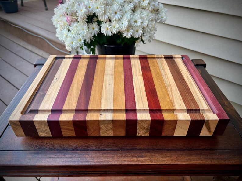 May include: A large, rectangular cutting board made of various types of wood. The board has a striped pattern of light and dark wood, with a darker wood border. The board is sitting on a wooden table.