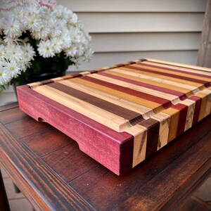 May include: A large, rectangular cutting board made of various types of wood. The board has a dark purple border and is striped with different shades of brown, red, and yellow wood.