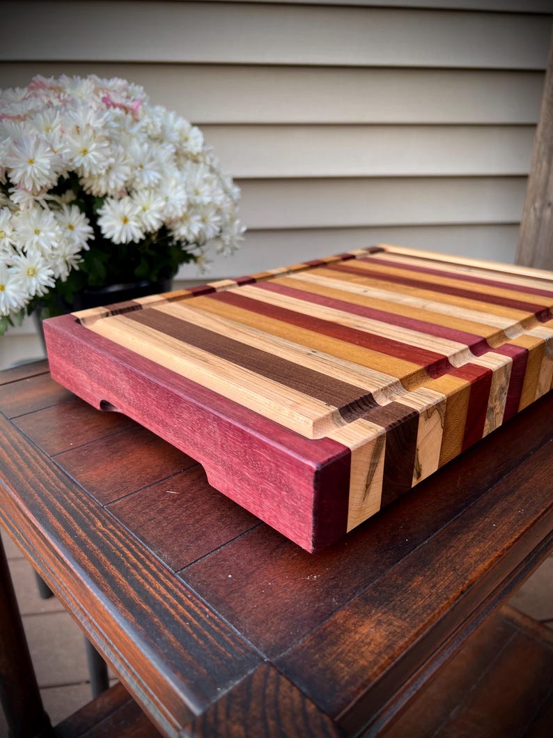 May include: A large, rectangular cutting board made of various types of wood. The board has a dark purple border and is striped with different shades of brown, red, and yellow wood.