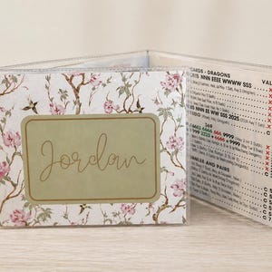 Puede incluir: Una cartera personalizada con un diseño floral con flores rosas y hojas verdes sobre un fondo blanco. El nombre "Jordan" se muestra en una etiqueta rectangular. La cartera tiene ranuras transparentes para tarjetas.