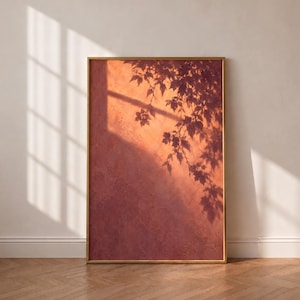 Könnte beinhalten: Ein rechteckiger Kunstdruck in einem dünnen goldenen Rahmen mit einer strukturierten, terrakottafarbenen Oberfläche. Das Bild zeigt einen scharfen, dunklen Schatten von belaubten Ästen, der auf die warm orangefarbene Wand geworfen wird.