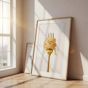 May include: A framed art print featuring a golden fork with a nest of pasta. The artwork is displayed on a light-colored wall, leaning against a second, smaller framed print. The room has a wooden floor and a large window.