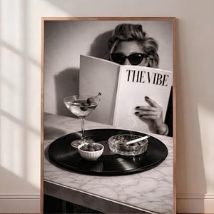 May include: A black and white wall art print featuring a person in sunglasses reading a magazine titled THE VIBE. On a table sits a vinyl record, a martini glass with olives, a small bowl of olives, and a glass ashtray with a lit cigarette.