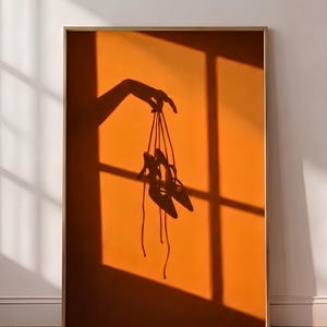 Könnte beinhalten: Ein gerahmter Wandkunstdruck mit einer dunklen Silhouette vor einem leuchtend orangefarbenen Hintergrund. Das Bild zeigt den Schatten einer Hand, die ein Paar hochhackige Schnürschuhe an ihren langen, dünnen Riemen hält, was einen Kontrast erzeugt.