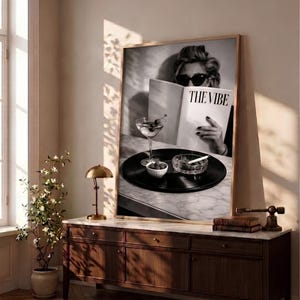 May include: A large framed black and white wall art print featuring a person wearing dark sunglasses while reading a magazine titled THE VIBE. A martini glass, a small bowl of olives, and a glass ashtray with a cigarette sit on a vinyl record.