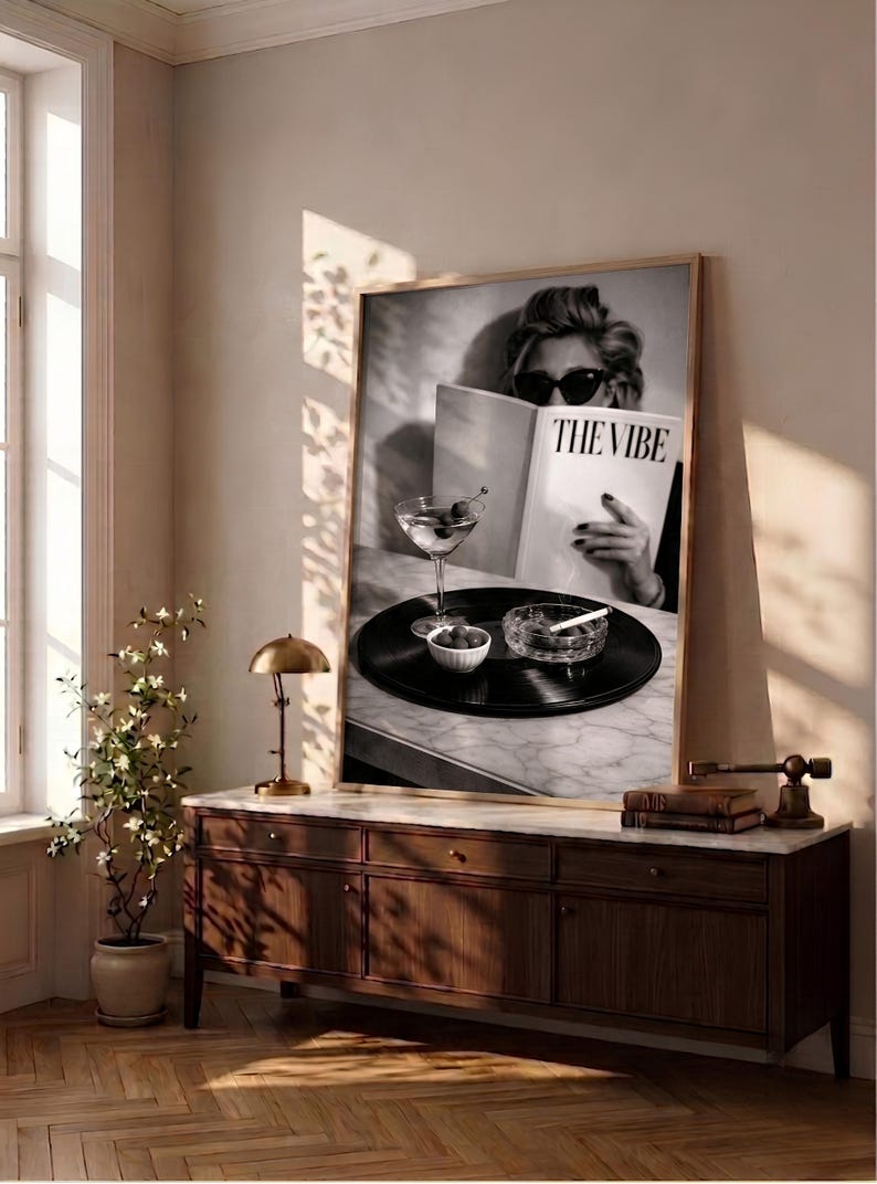 May include: A large framed black and white wall art print featuring a person wearing dark sunglasses while reading a magazine titled THE VIBE. A martini glass, a small bowl of olives, and a glass ashtray with a cigarette sit on a vinyl record.