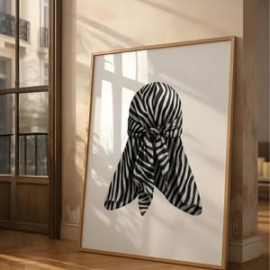 Puede incluir: Una impresión artística enmarcada con un durag a rayas de cebra en blanco y negro. El durag está atado en la parte posterior y cuelga hacia abajo. La obra de arte se muestra sobre un fondo blanco, con un marco de madera. La imagen está apoyada contra una pared en una habitación con pisos de madera.