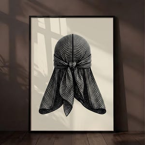 May include: A framed wall art print featuring a black durag covered in small, reflective silver studs. The durag is tied in the center with two long tails hanging down, displayed against a solid light beige background within a thin black frame.
