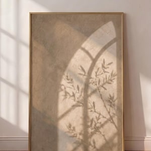Könnte beinhalten: Ein rechteckiger Wandkunstdruck in einem dünnen goldenen Rahmen mit strukturiertem beigen Hintergrund, auf den ein weicher, bogenförmiger Fensterschatten geworfen wird, überlagert von der Silhouette blättriger Zweige einer Pflanze.