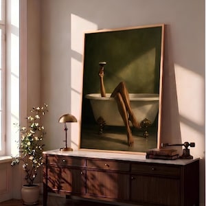 May include: A framed vertical wall art print depicting a person with legs extended out of a white clawfoot bathtub, holding a dark cocktail glass aloft. The background is a solid dark olive green, and the tub features ornate gold-toned feet.