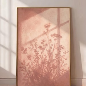 May include: A rectangular wall art print in a thin gold frame featuring a soft, dusty rose-colored background with a dark, silhouetted shadow of wildflowers and a window pane cast across the surface, creating a layered, textured visual effect.