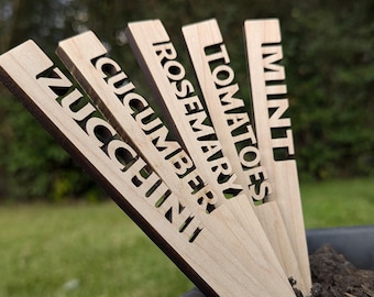 May include: Wooden garden markers with cut-out lettering. The markers are labeled "ZUCCHINI", "CUCUMBER", "ROSEMARY", "TOMATOES", and "MINT". The markers are light brown and are placed in a dark container with soil.