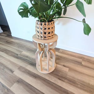 May include: A potted Monstera plant in a woven basket sits atop a light wood plant stand. The stand has a round top and a decorative base with curved, interlocking wooden pieces. The plant has large, green, split-leaf foliage.