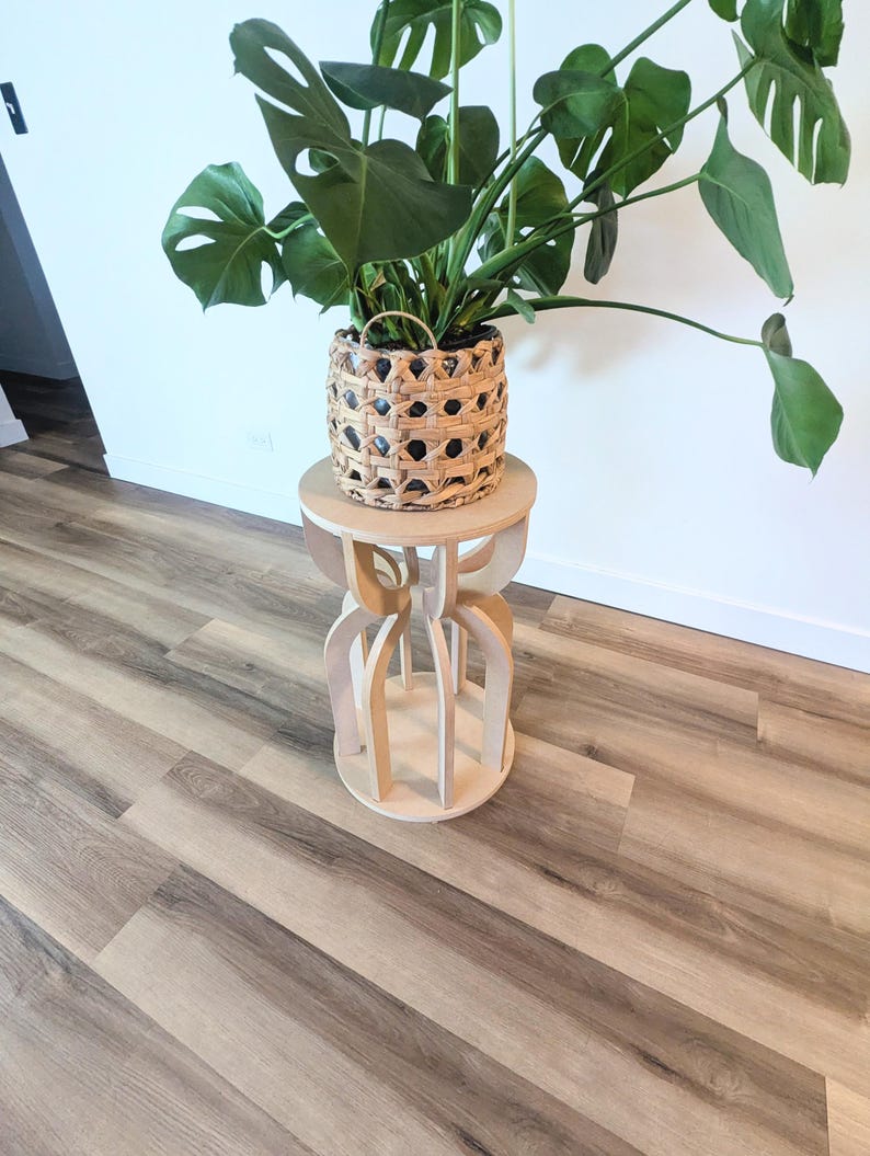 May include: A potted Monstera plant in a woven basket sits atop a light wood plant stand. The stand has a round top and a decorative base with curved, interlocking wooden pieces. The plant has large, green, split-leaf foliage.