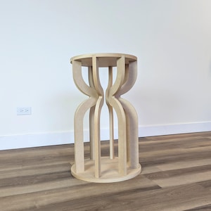 May include: A light-colored, wooden side table with a round top and a unique hourglass-shaped base. The base is constructed from curved, vertical supports that connect to a circular base. The table is approximately 24 inches tall.