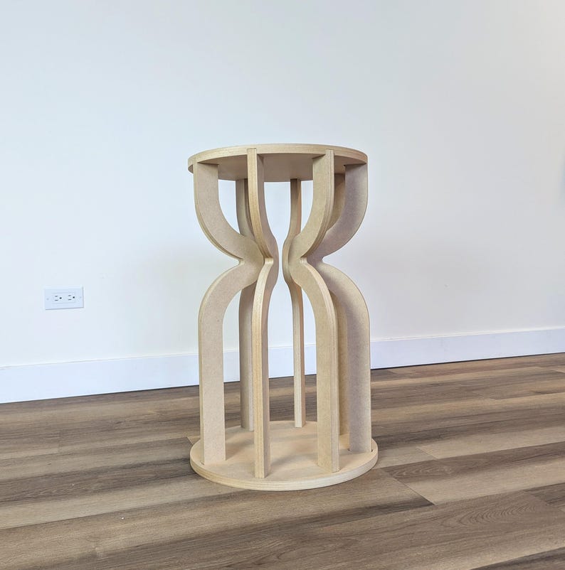 May include: A light-colored, wooden side table with a round top and a unique hourglass-shaped base. The base is constructed from curved, vertical supports that connect to a circular base. The table is approximately 24 inches tall.