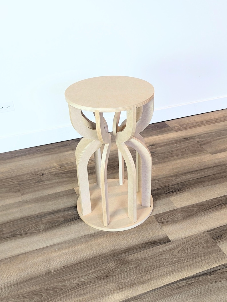 May include: A light-colored wooden stool with a round seat and a unique, open-legged base. The stool's design features curved supports that connect the seat to a circular base. The wood has a natural, unfinished appearance, and the stool is set against a white wall.