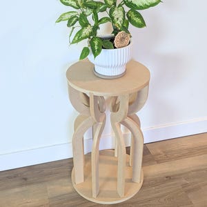 May include: A small, unfinished wooden plant stand with a round top and base, and a decorative, open-sided design. A white ceramic pot with a green and white plant sits on top. The stand is approximately 24 inches tall.