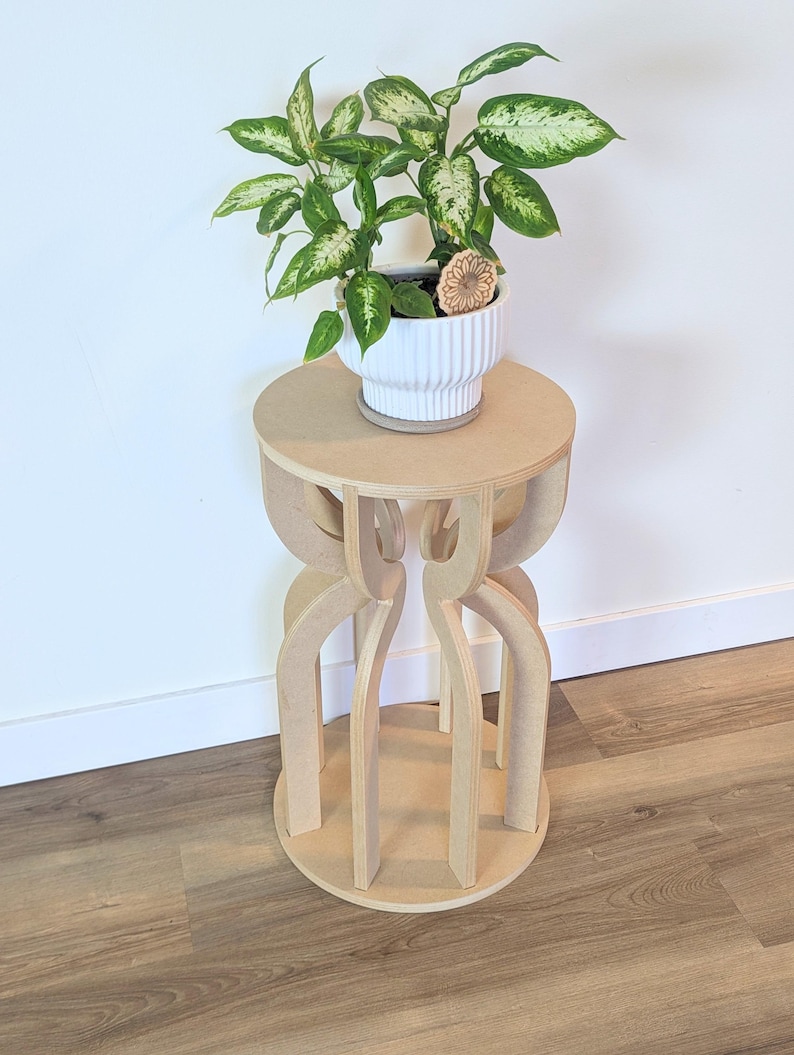 May include: A small, unfinished wooden plant stand with a round top and base, and a decorative, open-sided design. A white ceramic pot with a green and white plant sits on top. The stand is approximately 24 inches tall.